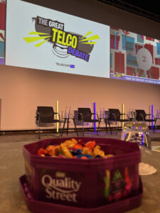 A picture of a tin of quality street in the foreground, with the Great Telco Debate logo in the background. 